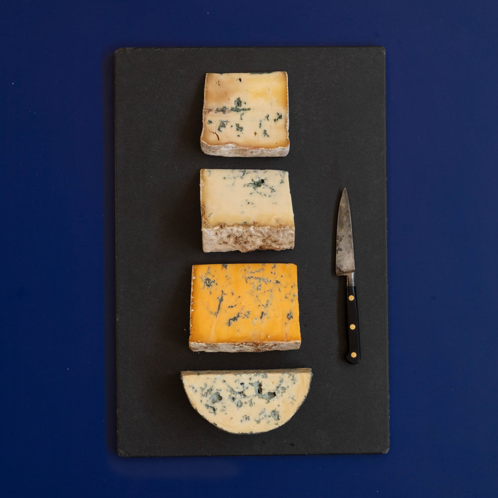 Four wedges and slices of assorted blue cheeses arranged vertically on a dark gray slate serving board, with a small cheese knife on the right, against a solid dark blue background.
