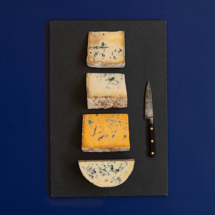 Four wedges and slices of assorted blue cheeses arranged vertically on a dark gray slate serving board, with a small cheese knife on the right, against a solid dark blue background.