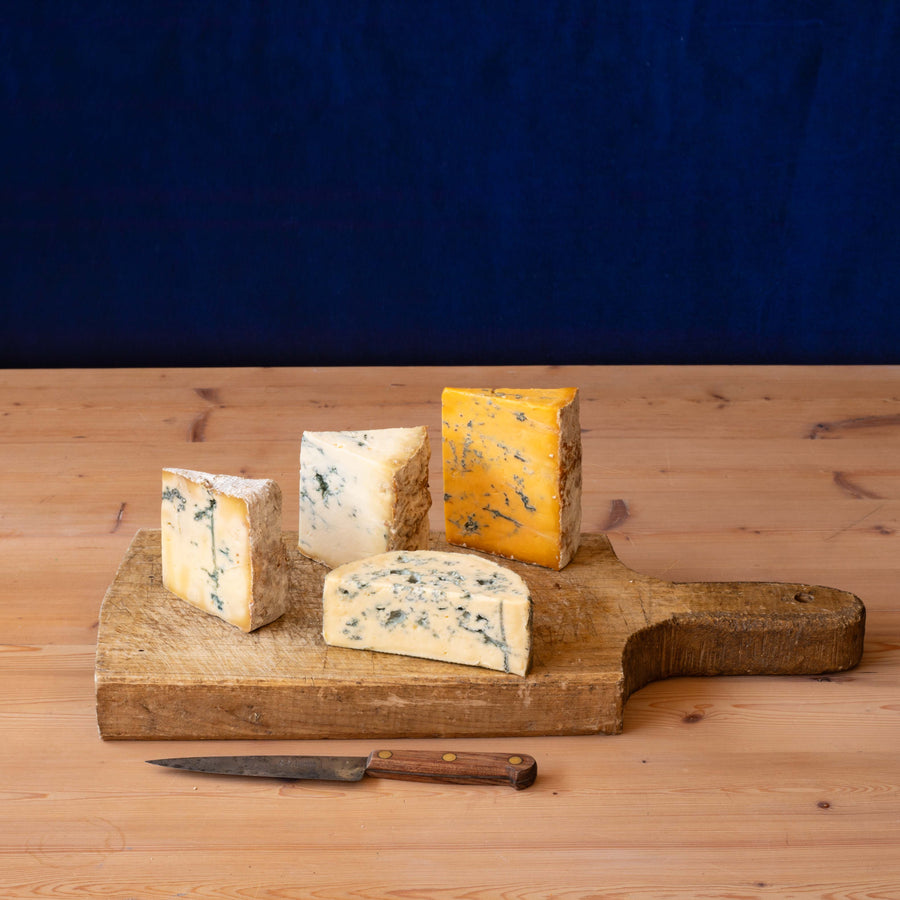 Four wedges and slices of assorted cheeses displayed on a rustic wooden cutting board, with a small knife in front, set on a light wood tabletop against a dark blue backdrop.