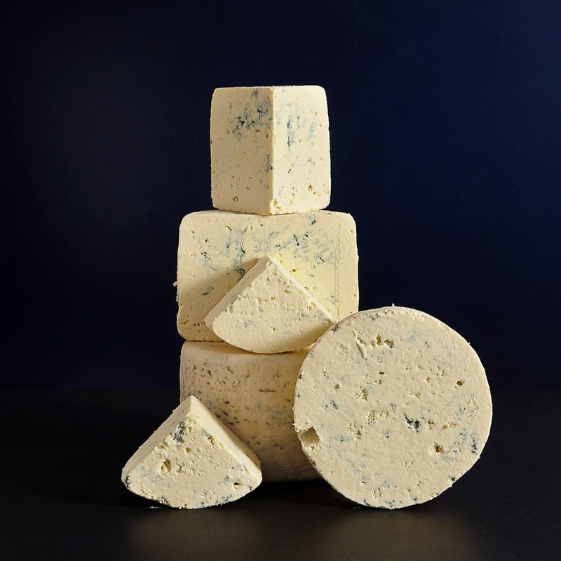 Tower of a whole and cut pieces of Devon Blue vegetarian cow’s milk cheese, showing the delicate blue veining and crumbly, buttery texture